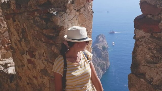 A woman tourist with a backpack and a hat sits on the fortress wall in the fortress of Alanya, Turkey. Beautiful sea landscape with a fortress.