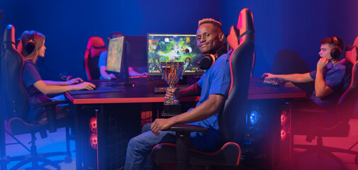Fototapeta na wymiar Streamer young african man rejoices in victory professional gamer playing online games computer with headphones, neon colour, soft focus