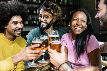 Happy multiracial young friends having fun together drinking beer at brewery bar - Millennial people celebrating toasting alcohol drinks at restaurant