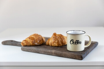 Two croissants, a cup with coffee inscription on a wooden board on a white table. Place for text
