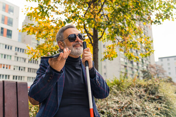 Elegant blind bearded grey-haired mature man with dark sunglasses on holding a walking stick,...