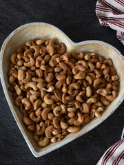 Roasted cashew nuts on a heart-shaped plate on a dark background. Vertical shot. Top table view, a close-up.