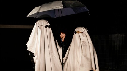 Couple of people wearing ghost costumes and holding umbrella in dark street at night