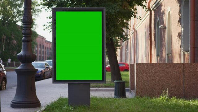 4k Green mockup billboard stands on street in city spbd. Closeup view of vertical light display is standing by road, cars driving and people walking along sidewalk in town on summer day. Large