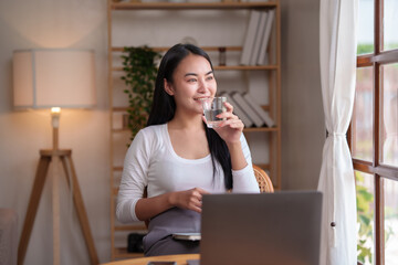 Beautiful Asian woman sitting and drinking water to relax from working on her laptop on the computer to reduce the fatigue caused by work at home.