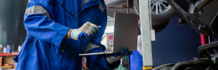 Young man is mechanic using laptop computer checking engine of car in the garage, worker examining and diagnostic automobile, technician and maintenance of vehicle, transport concept.