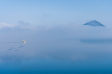Fototapeta na wymiar 幻想的な霧の中に浮かび上がる山と湖を進む白い船