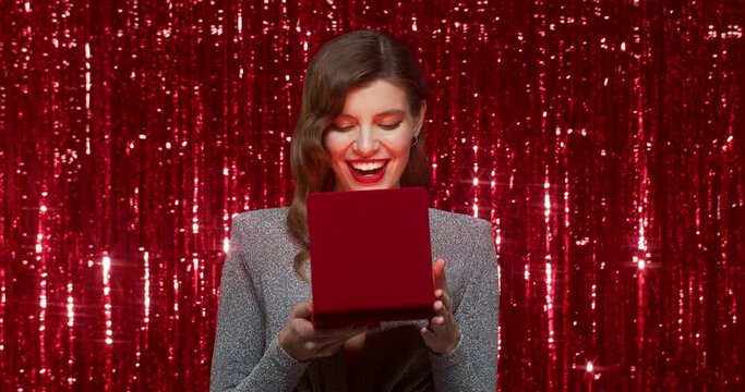 Surprised woman opening opening luxury red necklace box with christmas gift with magic shining light on red tinsel background