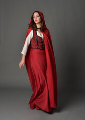 Fototapeta premium Full length portrait of beautiful brunette woman wearing red medieval fantasy costume with long skirt and flowing hooded cloak. Standing pose with gestural hand poses, isolated on grey studio backgrou