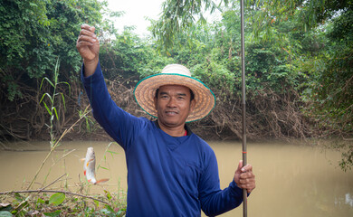 A middle-aged Asian man, using a basic fishing rod, fishes at the river and shows it up in front of the camera. with a smile and looking at the camera