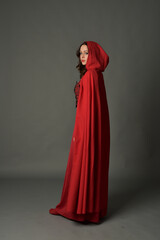 Naklejka premium Full length portrait of woman wearing red medieval fantasy costume, flowing hooded cloak. Standing pose in backview, gestural hand poses, walking away from camera isolated on grey studio background.