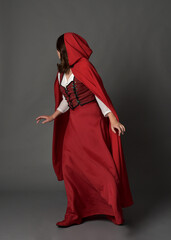 Full length portrait of woman wearing red medieval fantasy costume, flowing hooded cloak. Standing pose in backview, gestural hand poses, walking away from camera isolated on grey studio background.