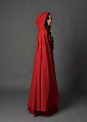 Naklejka premium Full length portrait of woman wearing red medieval fantasy costume, flowing hooded cloak. Standing pose side profile, gestural hand poses, walking away from camera isolated on grey studio background.