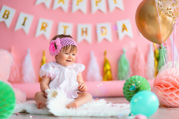 Smiling baby girl sits near paper balls and balloons in holiday dress on pink birthday background