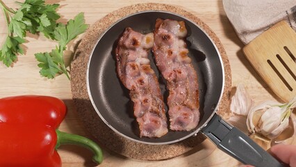 The cook puts on a served table a frying pan with fried eggs and bacon. On the table are greens and red pepper. European breakfast or lunch concept