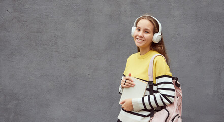 cheerful girl student with a pink backpack in headphones holds a textbook against a gray wall