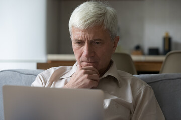 Fototapeta premium Thoughtful grey-haired mature man learning laptop app or software, looking pensive and serious sit on sofa at home, close-up. Older people using modern tech, experiences difficulties with technology