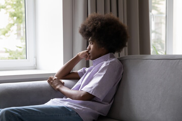 Sad African insecure teenage guy outcast sit on sofa staring through window, looks pensive thinks...