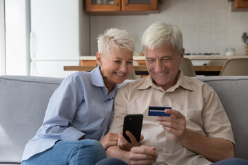 Happy older couple sit on sofa at home enjoy on-line purchase using smartphone and credit card. Mature spouses do electronic shopping, secure payment, make instant money transfer, ecommerce clients