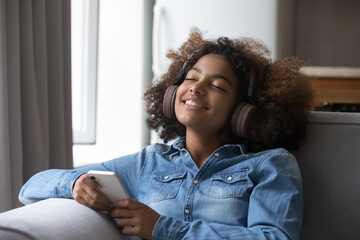 Peaceful African girl holds mobile phone listens music via wireless headphones with eyes closed leaned on sofa at home, teenager enjoy leisure, relish favourite song spend time indoors feel carefree