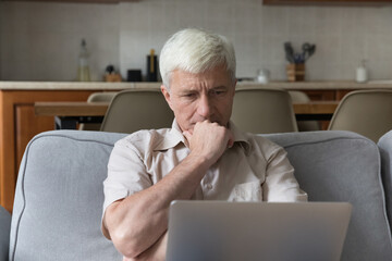 Serious thoughtful older man sit on couch with laptop, learns computer software or new program,...