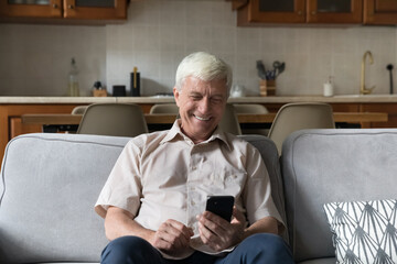 Senior man looking at cellphone screen, watch funny videos, have fun using new mobile application spend time on internet sit on couch at home. Older person and modern technology usage, leisure online