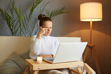 Portrait of tired sad sleepy woman wearing white sweater sitting on sofa and working on notebook,...