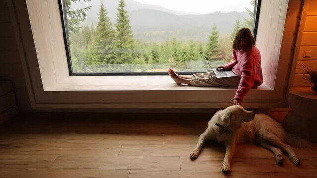 Woman works on laptop while sitting on a window with great view on mountains. Her dog lying nearby. Remote work, escape to nature and home coziness concept