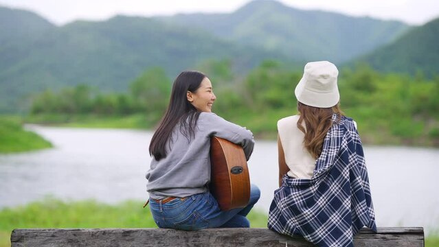 Happy Asian woman friends enjoy and fun outdoor lifestyle camping together in forest mountain on summer travel vacation. Attractive girl singing and playing guitar by the lake near the camp at sunset