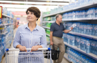 Mature woman considering bottle of vermouth in supermarket - obrazy, fototapety, plakaty