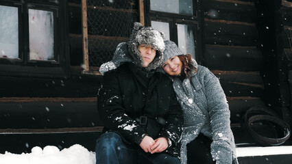 Obraz na płótnie Canvas winter portrait. Happy, funny couple in love, man and woman, dressed in warm winter clothes, fully covered with snowflakes, are sitting on bench by an old wooden house and hugging. happy time on snowy