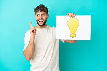 Young handsome caucasian man isolated on blue background holding a placard with bulb icon and...
