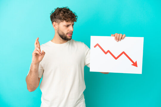 Young handsome caucasian man isolated on blue background holding a sign with a decreasing statistics arrow symbol with fingers crossing