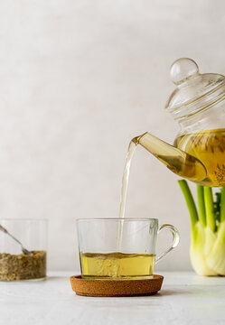 Fennel tea in a glass cup, fresh fennel bulb, seeds and tea pot pouring tea to cup. White wooden table with light background
