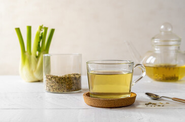 Fennel tea in a glass cup, fresh fennel bulb, seeds in jar, tea pot on white wooden table with light background