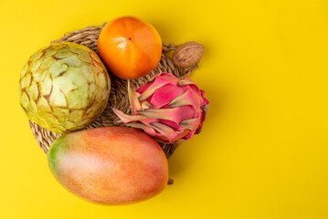 Autumn tropical fruits,mango,custard apple,pitahaya and persimmon, on a shallow plate of cattail with a yellow background,copy space.