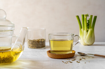 Fennel tea in a glass cup, fresh fennel bulb, seeds in jar, tea pot on white wooden table with...