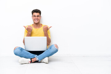Young man sitting on the floor isolated on white background with thumbs up gesture and smiling