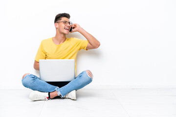 Young man sitting on the floor isolated on white background keeping a conversation with the mobile phone