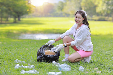 Young beautiful asian volunteer woman hold garbage bag and picking up litter while cleaning plastic bottle from lawn and staring at camera during a volunteering environmental cleanup area park event
