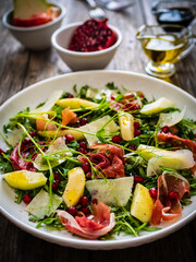 Fresh salad - prosciutto di Parma, pomegranate, pear, leafy vegetables and parmesan on wooden background
