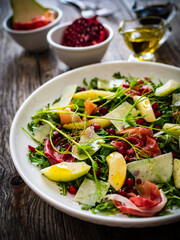 Fresh salad - prosciutto di Parma, pomegranate, pear, leafy vegetables and parmesan on wooden background
