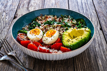 Boiled eggs with avocado, boiled red and white rice, cherry tomatoes, spinach and parmesan on wooden table
