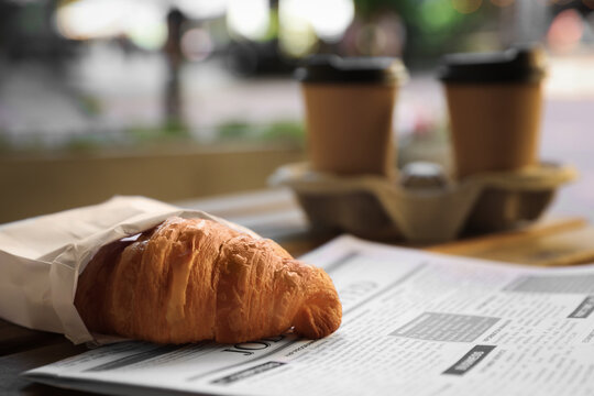 Tasty croissant, newspaper and paper cups of coffee on wooden table outdoors