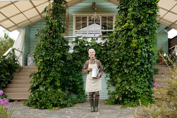 Happy old woman in workwear standing in front of camera against cottage or summer house overgrown with green grape leaves © pressmaster
