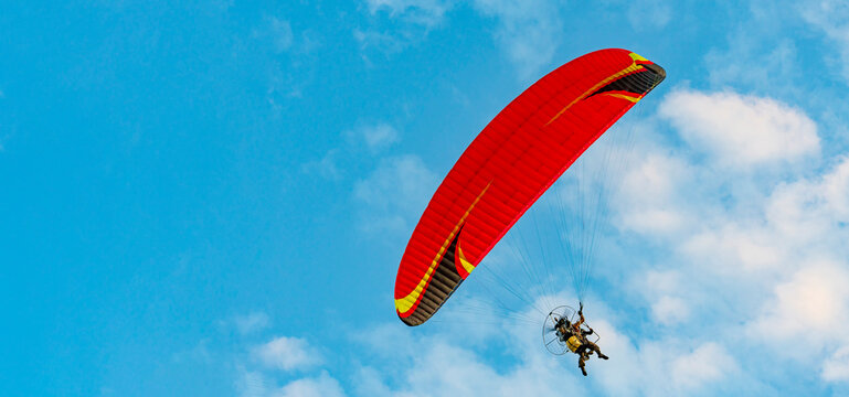 Against background of blue summer cloudy sky, tandem of paragliders flies on red ultralight aircraft - paraglider. Extreme sport and recreation.