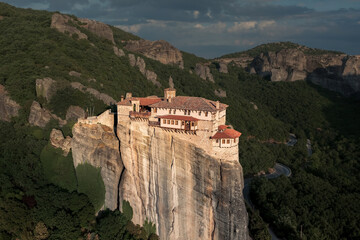 Fototapeta na wymiar Brightly lit Christian monastery on cliff surrounded by mountain cliffs at sunset, Meteora, Greece