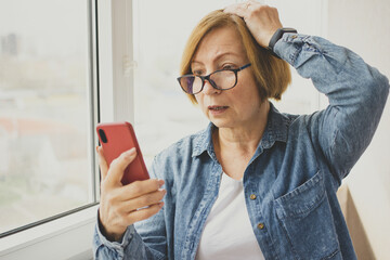 Worried mature woman looking at smartphone with amazement open mouth, reading disturbing news in...