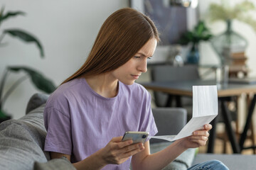 Worried young woman  reading paper bill pay online using mobile phone at home. Female holding bank...