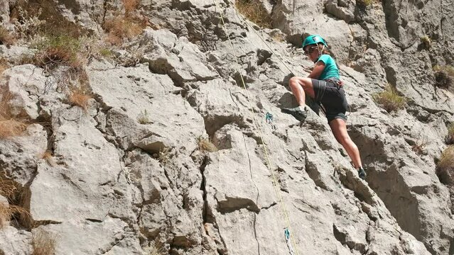 Woman in protective helmet and shoes climbing on vertical cliff rock wall using top rope and climbing harness in Paklenica National park site, Croatia. Active extreme sports time spending 4K concept.